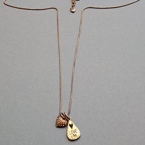 Puede incluir: Un delicado collar con dos colgantes. Un colgante es un peque&ntilde;o dije en forma de coraz&oacute;n cubierto de peque&ntilde;as piedras transparentes. El otro colgante tiene forma de l&aacute;grima con la palabra "LOVE" y un dise&ntilde;o de coraz&oacute;n. La cadena es fina y plateada.