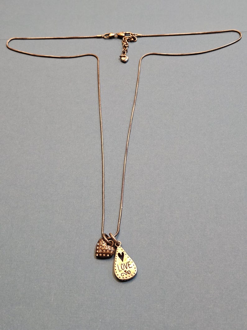 Puede incluir: Un delicado collar con dos colgantes. Un colgante es un peque&ntilde;o dije en forma de coraz&oacute;n cubierto de peque&ntilde;as piedras transparentes. El otro colgante tiene forma de l&aacute;grima con la palabra "LOVE" y un dise&ntilde;o de coraz&oacute;n. La cadena es fina y plateada.