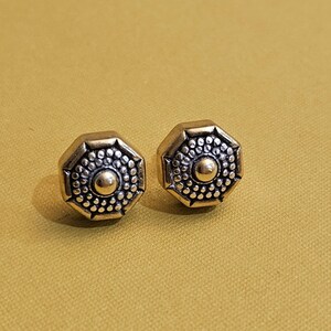 May include: A pair of octagonal stud earrings with a vintage aesthetic. Each earring features a central gold-colored sphere surrounded by a detailed pattern of small dots and an outer frame. The earrings are set against a mustard-yellow background.