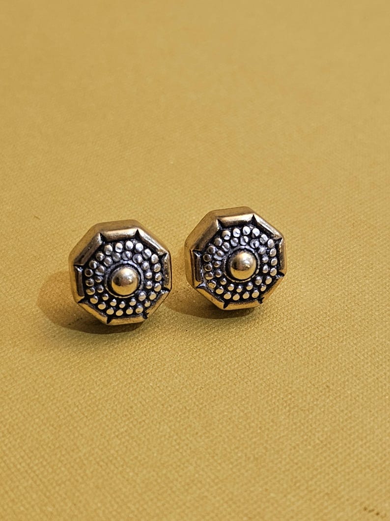 May include: A pair of octagonal stud earrings with a vintage aesthetic. Each earring features a central gold-colored sphere surrounded by a detailed pattern of small dots and an outer frame. The earrings are set against a mustard-yellow background.