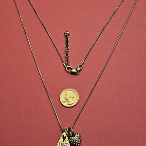 Puede incluir: Un collar plateado con un colgante en forma de l&aacute;grima grabado con la palabra "LOVE" y un colgante en forma de coraz&oacute;n. El collar se muestra sobre un fondo rojo junto a una moneda.