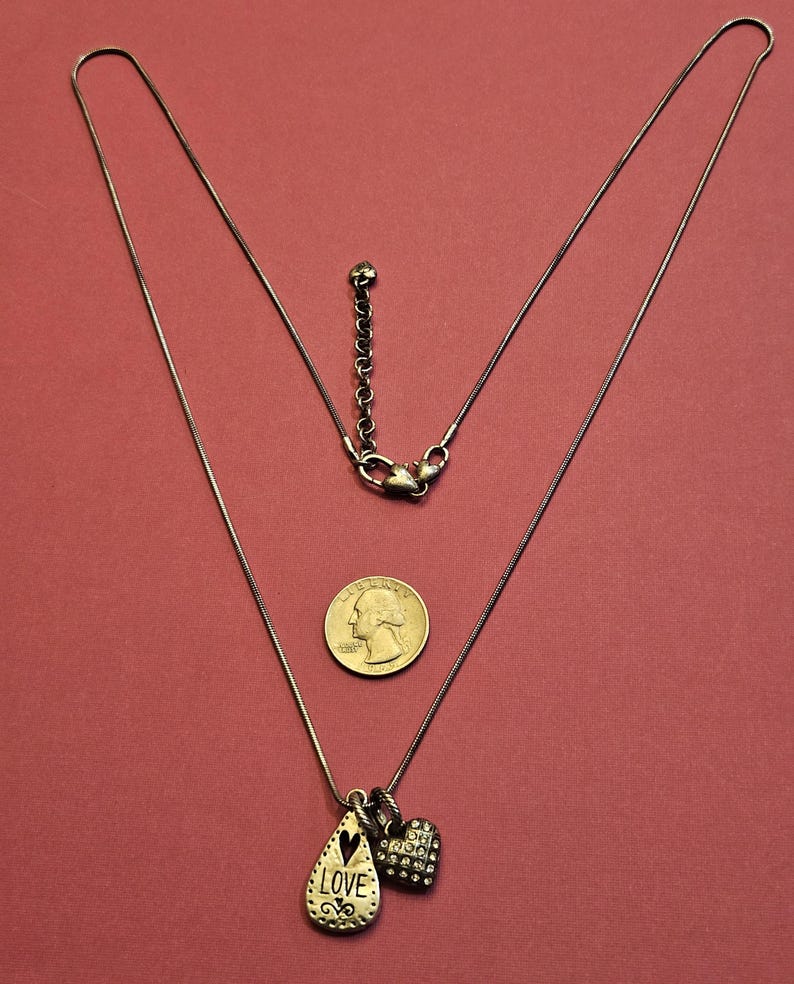 Puede incluir: Un collar plateado con un colgante en forma de l&aacute;grima grabado con la palabra "LOVE" y un colgante en forma de coraz&oacute;n. El collar se muestra sobre un fondo rojo junto a una moneda.