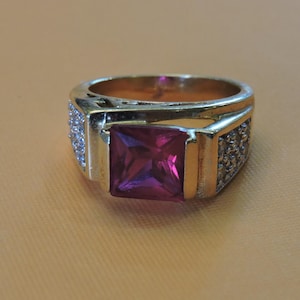 May include: A gold-toned ring with a large, square, magenta-colored gemstone at the center. The ring's band has small, clear stones on each side of the central stone. The ring is set against a light brown background.