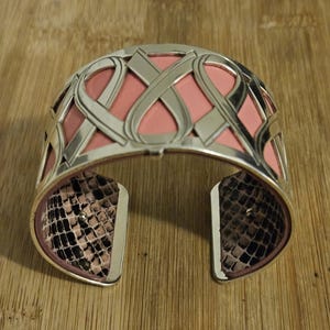 May include: Silver-tone cuff bracelet with a pink interior and a snakeskin pattern on the inside of the opening. The bracelet features an intricate, openwork design with interwoven shapes.