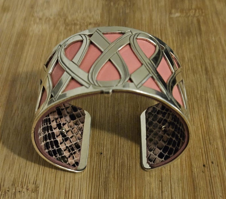 May include: Silver-tone cuff bracelet with a pink interior and a snakeskin pattern on the inside of the opening. The bracelet features an intricate, openwork design with interwoven shapes.