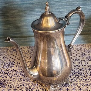 Op de afbeelding: Een zilverkleurige, antieke koffiepot met een gebogen tuit en een sierlijke handgreep. De pot heeft een deksel met een decoratieve knop en staat op een patroon oppervlak. Het ontwerp is klassiek en elegant.