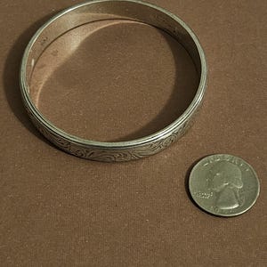 May include: A silver-colored bangle bracelet with an engraved floral pattern. The bracelet is circular and sits next to a U.S. quarter coin. The bracelet's diameter is approximately 2.5 inches.