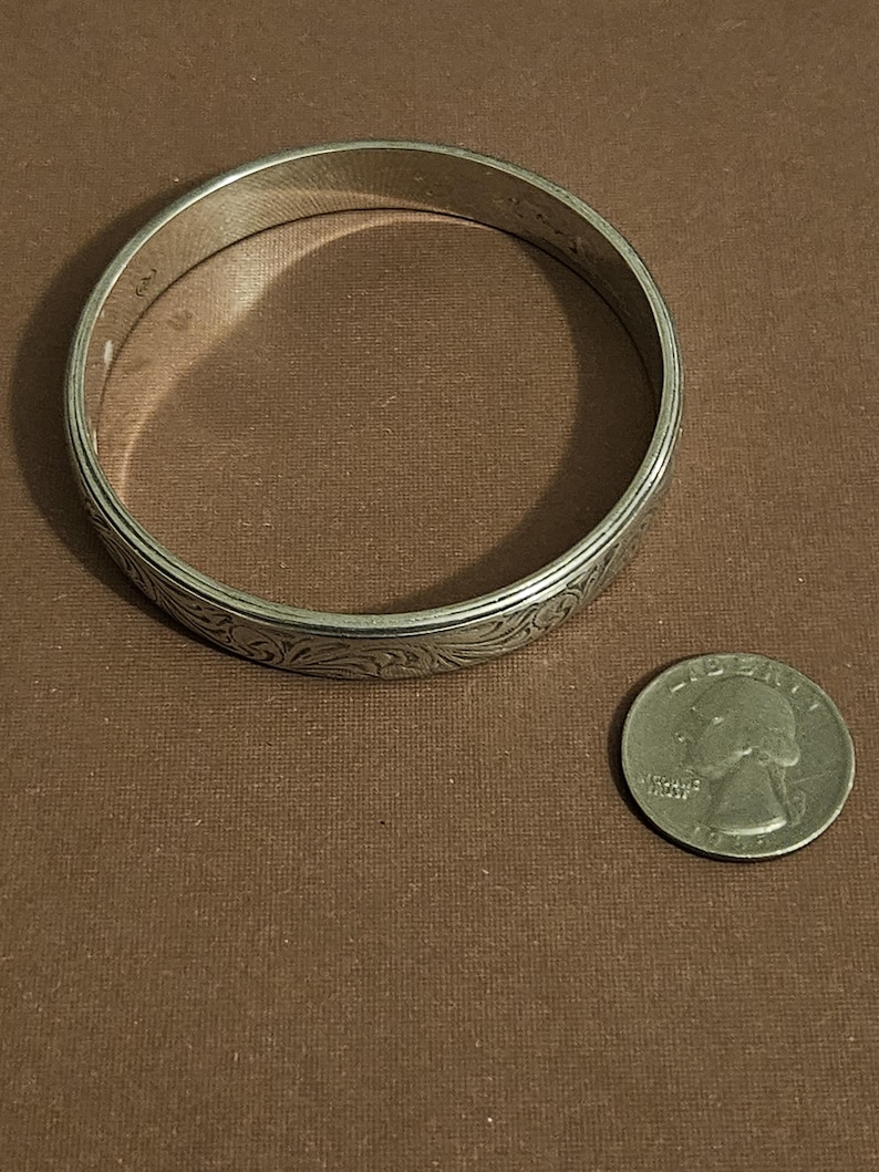 May include: A silver-colored bangle bracelet with an engraved floral pattern. The bracelet is circular and sits next to a U.S. quarter coin. The bracelet's diameter is approximately 2.5 inches.