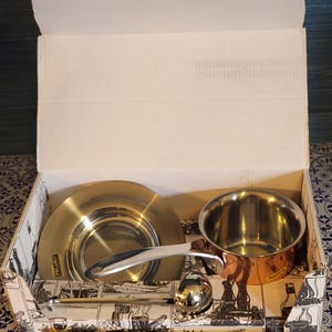 May include: A stainless steel and copper cookware set inside a cardboard box. The set includes a small saucepan, a serving spoon, a ladle, and a lid. The box has a black and white illustration on the sides.