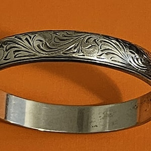 May include: Silver-colored bangle bracelet with an engraved floral pattern. The bracelet has a polished, smooth interior and a detailed, textured exterior. The engraved design features swirling leaves and vines. The bracelet is a classic piece of jewelry.