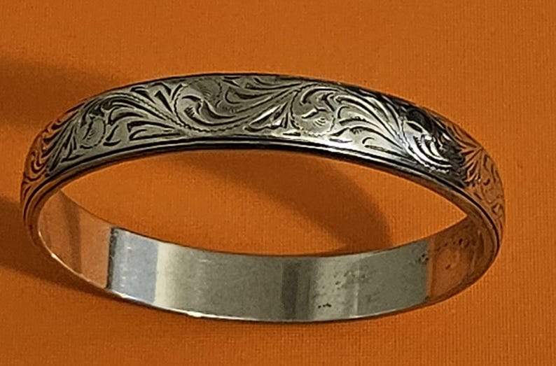 May include: Silver-colored bangle bracelet with an engraved floral pattern. The bracelet has a polished, smooth interior and a detailed, textured exterior. The engraved design features swirling leaves and vines. The bracelet is a classic piece of jewelry.