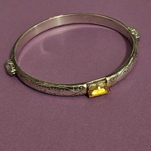 May include: Silver-toned hinged bangle bracelet with engraved floral patterns. The bracelet features a rectangular, yellow gemstone clasp and two smaller, round gemstones. The bracelet is displayed on a purple surface.