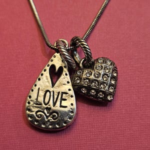 Puede incluir: Un collar plateado con dos colgantes. Uno es en forma de l&aacute;grima con la palabra "LOVE" y un coraz&oacute;n recortado. El otro es en forma de coraz&oacute;n, incrustado con peque&ntilde;as piedras transparentes. El collar se muestra sobre un fondo rosa.