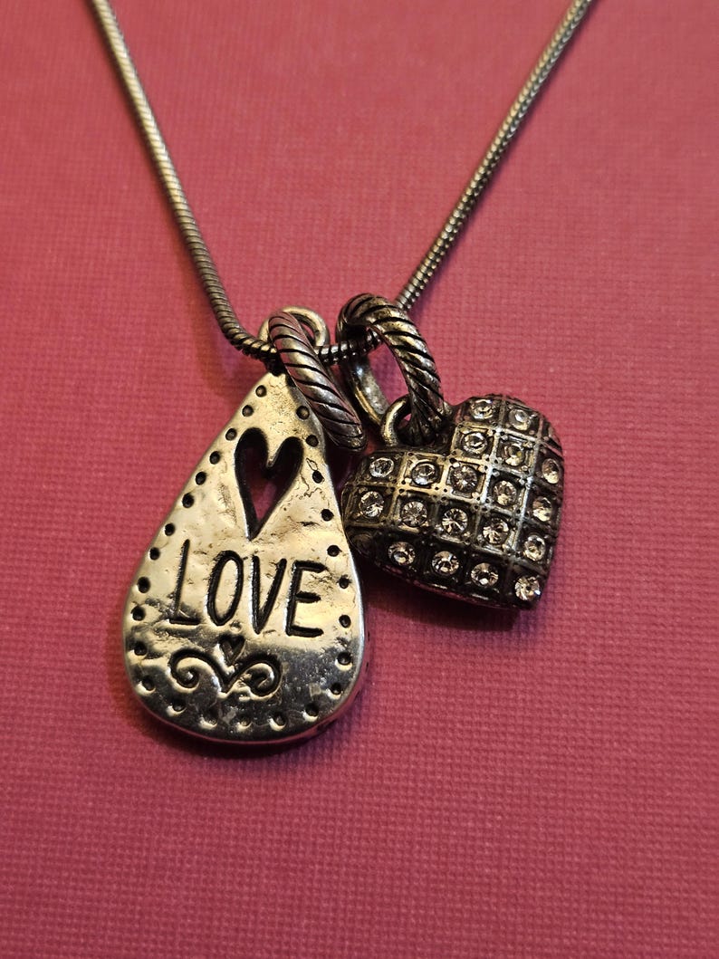 Puede incluir: Un collar plateado con dos colgantes. Uno es en forma de l&aacute;grima con la palabra "LOVE" y un coraz&oacute;n recortado. El otro es en forma de coraz&oacute;n, incrustado con peque&ntilde;as piedras transparentes. El collar se muestra sobre un fondo rosa.