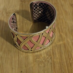 May include: A silver cuff bracelet with a pink leather interior and a snakeskin pattern on the inside edges. The exterior features a decorative, openwork design with pink leather accents.