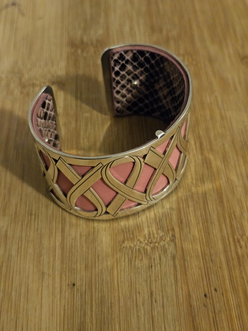 May include: A silver cuff bracelet with a pink leather interior and a snakeskin pattern on the inside edges. The exterior features a decorative, openwork design with pink leather accents.