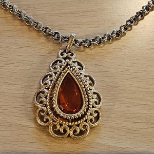 May include: A vintage pendant necklace featuring a large, teardrop-shaped, amber-colored stone set in a gold-toned filigree frame. The pendant hangs from a silver-toned chain. The necklace is displayed on a light brown wooden surface.