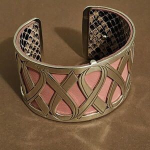 May include: A wide cuff bracelet with a pink leather interior and a silver-tone metal exterior. The metal is designed with an interwoven ribbon pattern. The interior of the bracelet's ends features a snakeskin pattern.