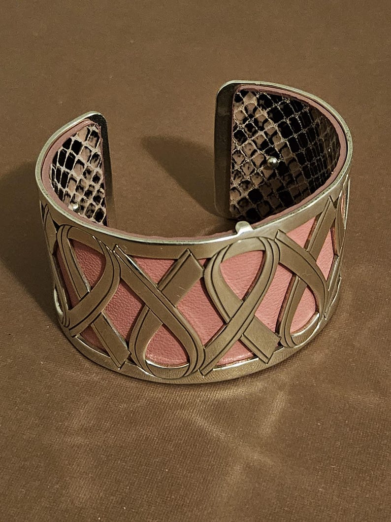 May include: A wide cuff bracelet with a pink leather interior and a silver-tone metal exterior. The metal is designed with an interwoven ribbon pattern. The interior of the bracelet's ends features a snakeskin pattern.