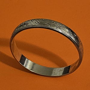 May include: A silver bangle bracelet with an engraved floral pattern. The bracelet is circular and has a polished finish. The engraved design is visible on the top surface of the bracelet.