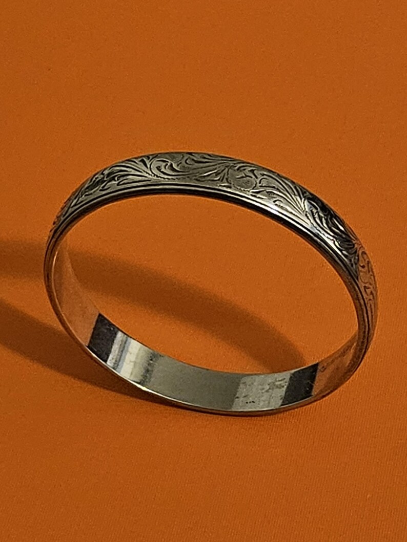 May include: A silver bangle bracelet with an engraved floral pattern. The bracelet is circular and has a polished finish. The engraved design is visible on the top surface of the bracelet.