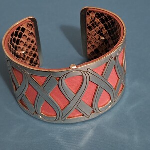 May include: Silver-tone cuff bracelet with a pink leather interior and a gray ribbon design. The interior edges feature a snakeskin pattern. A stylish accessory for everyday wear.