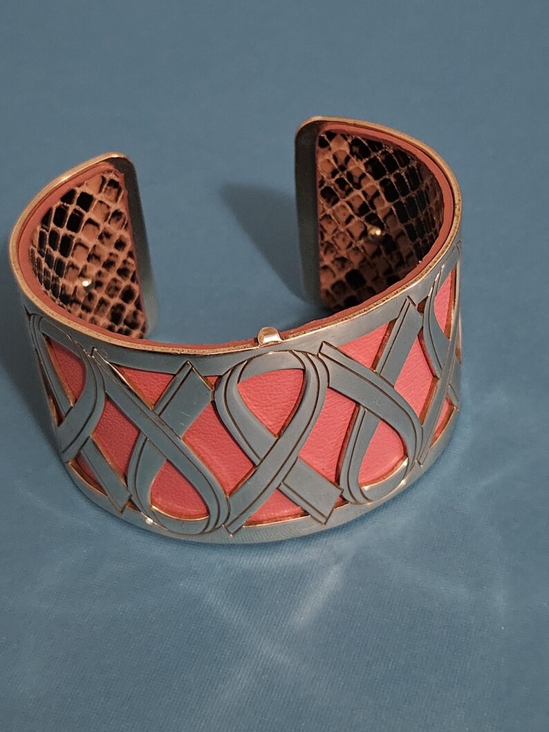 May include: Silver-tone cuff bracelet with a pink leather interior and a gray ribbon design. The interior edges feature a snakeskin pattern. A stylish accessory for everyday wear.