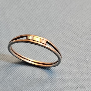 May include: A copper-toned ring with a rectangular inlay of various colors. The ring has a split band design, with a thin band on either side of the inlay. The inlay features shades of white, brown, and yellow.