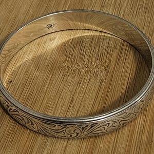 May include: A silver bangle bracelet with an engraved floral pattern. The bracelet has a smooth, polished finish and a delicate design. The bangle is circular and appears to be made of metal. The bracelet is resting on a wooden surface.