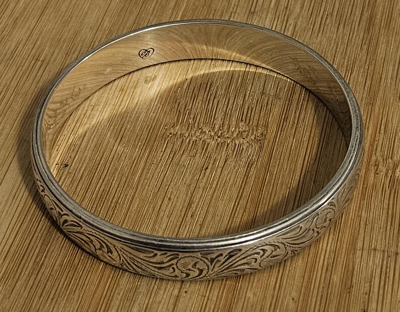 May include: A silver bangle bracelet with an engraved floral pattern. The bracelet has a smooth, polished finish and a delicate design. The bangle is circular and appears to be made of metal. The bracelet is resting on a wooden surface.