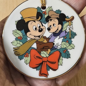 May include: A round Christmas ornament featuring Mickey Mouse and a friend, surrounded by holly and berries. Mickey wears a yellow hat, scarf, and brown gloves. A large red bow is at the bottom. The ornament has a gold trim and a gold cord for hanging.