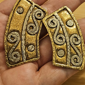 May include: A pair of large, gold and silver earrings with a Celtic knot design. The earrings are made of metal and have a textured finish.
