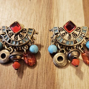 May include: A pair of gold tone dangle earrings with red and blue stones. The earrings feature a geometric design with a crescent moon shape and multiple dangling charms.