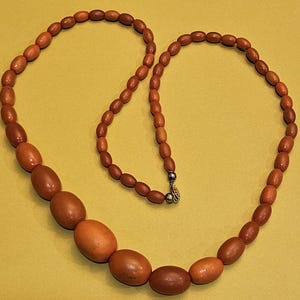 May include: A necklace featuring oval-shaped wooden beads in varying shades of brown and orange. The beads are strung together, with a silver-colored clasp. The necklace is displayed against a yellow-green background.