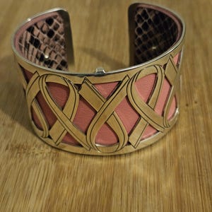 May include: A silver-toned cuff bracelet with a pink leather interior and a gold-colored, interwoven design. The interior features a snakeskin pattern. The bracelet is open at the top and bottom.