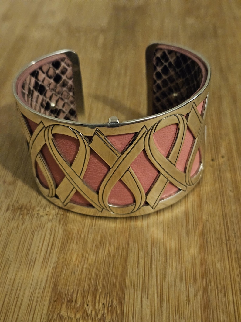 May include: A silver-toned cuff bracelet with a pink leather interior and a gold-colored, interwoven design. The interior features a snakeskin pattern. The bracelet is open at the top and bottom.