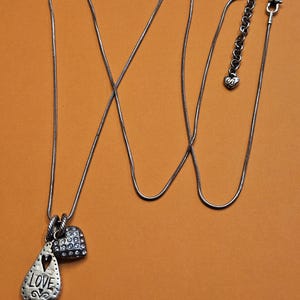 Puede incluir: Collar plateado con tres colgantes. Un colgante tiene forma de l&aacute;grima con la palabra "LOVE" y un coraz&oacute;n recortado. El segundo colgante es un peque&ntilde;o coraz&oacute;n cubierto de peque&ntilde;as piedras transparentes. El collar tiene una cadena ajustable.