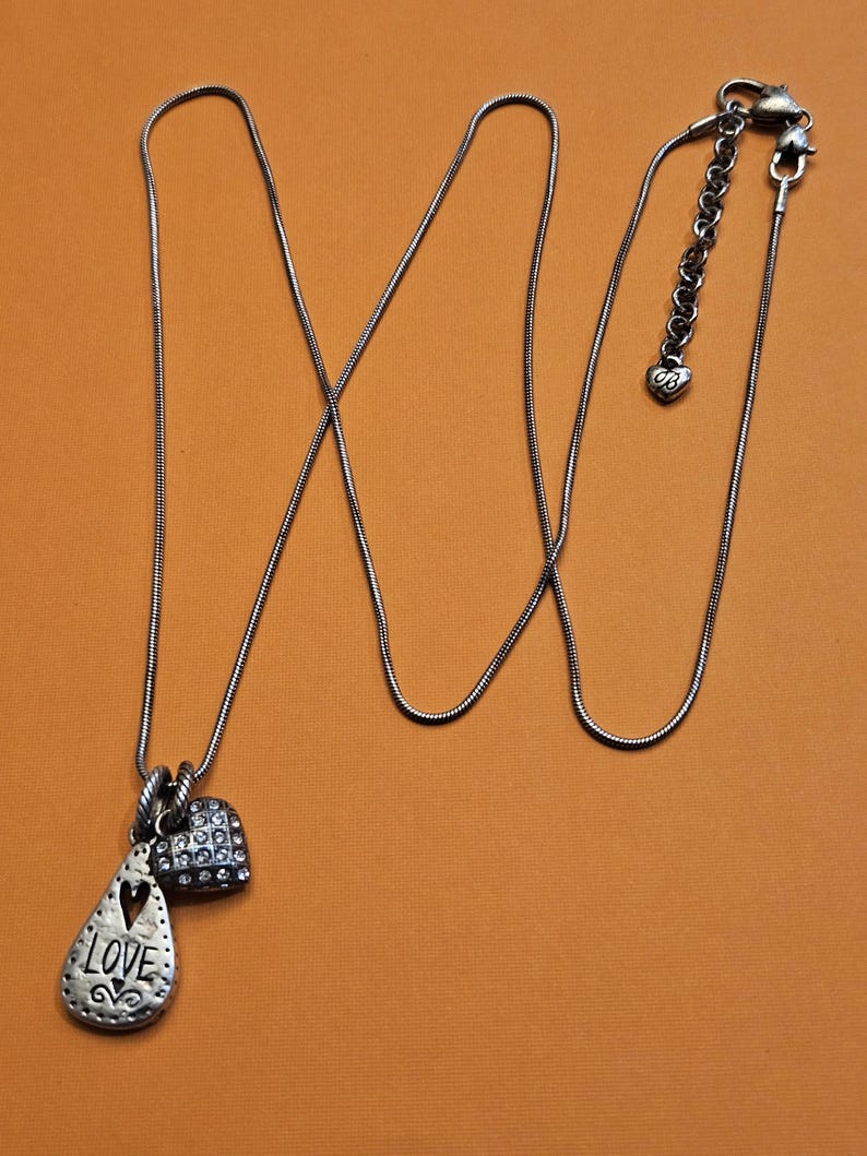 Puede incluir: Collar plateado con tres colgantes. Un colgante tiene forma de l&aacute;grima con la palabra "LOVE" y un coraz&oacute;n recortado. El segundo colgante es un peque&ntilde;o coraz&oacute;n cubierto de peque&ntilde;as piedras transparentes. El collar tiene una cadena ajustable.