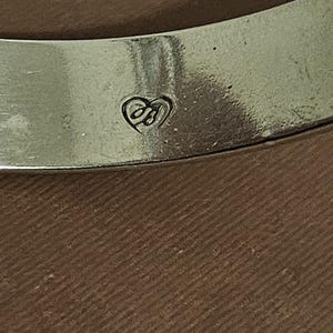 May include: Close-up of a silver-toned bangle bracelet. The bracelet has a heart-shaped logo with the initials "JB" engraved on the surface. The background is a textured brown surface.