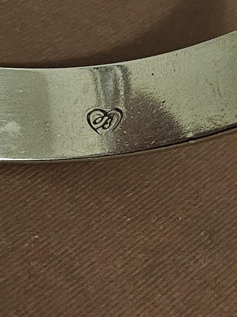 May include: Close-up of a silver-toned bangle bracelet. The bracelet has a heart-shaped logo with the initials "JB" engraved on the surface. The background is a textured brown surface.