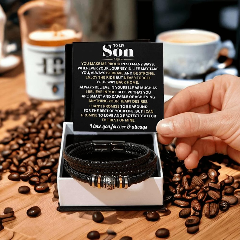May include: A black leather bracelet set in a white gift box, with a card that reads "To My Son" and a heartfelt message. The bracelet features silver and gold-toned accents. Coffee beans and a cup of coffee are in the background.