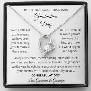 May include: A silver necklace with a heart-shaped pendant, featuring a central clear gemstone. The pendant is on a chain, presented in a gift box with text that reads "To Our Granddaughter On Your Graduation Day."
