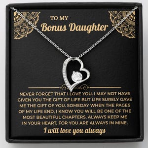 Peut inclure: Un collier avec pendentif en forme de cœur argenté et une pierre centrale en zircone cubique, présenté dans une boîte noire avec le texte doré TO MY Bonus Daughter et un message affectueux sur la vie et l'amour.
