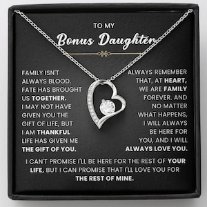 May include: A silver necklace with a heart-shaped pendant, featuring a central clear gemstone. The pendant hangs on a delicate chain, presented on a black background with text that reads "To My Bonus Daughter."