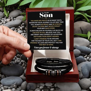 May include: A black leather bracelet with silver and copper accents is displayed inside a red wooden box. The box also contains a card with the text "TO MY SON" and a heartfelt message. The bracelet is a thoughtful gift.