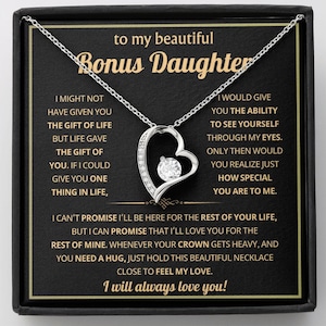 Puede incluir: Un collar plateado con un colgante en forma de corazón, con una piedra transparente central. El collar se presenta en una caja negra con el texto "to my beautiful Bonus Daughter" y un mensaje de amor.