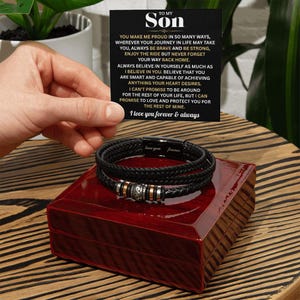 May include: Black leather bracelet with silver and gold accents, displayed on a red wooden box. A card with the text "TO MY SON" and a heartfelt message is held nearby. The bracelet is a thoughtful gift.