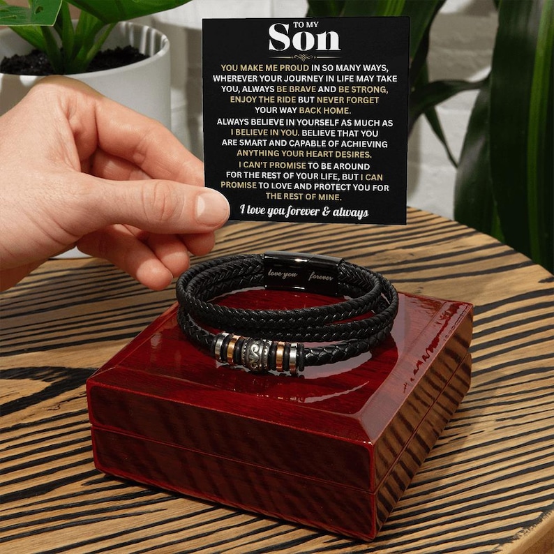 May include: Black leather bracelet with silver and gold accents, displayed on a red wooden box. A card with the text "TO MY SON" and a heartfelt message is held nearby. The bracelet is a thoughtful gift.