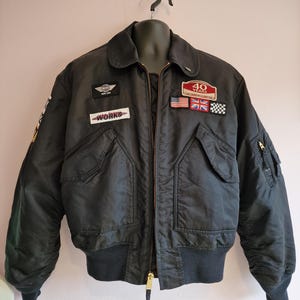 Puede incluir: Chaqueta bomber negra con múltiples parches, incluyendo "WORKS" y una insignia de 40 aniversario. Cuenta con cremallera frontal, dos bolsillos delanteros y dobladillo y puños acanalados. La chaqueta se exhibe en un maniquí.