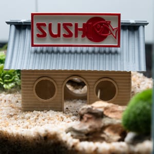 May include: A miniature sushi restaurant model with a gray roof and a tan wooden facade. The sign reads "SUSHI" in red letters with a fish graphic. The model has three circular openings and is set in a miniature aquarium environment.